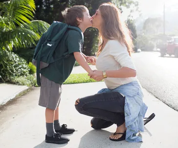 A mother kneels and kisses her young son goodbye as he heads to school with a backpack.