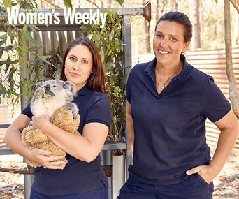 Two women in navy shirts, one holding a koala, in a natural setting. Appears from Australian Women's Weekly.