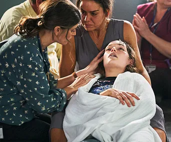 Three concerned people surround an unconscious woman wrapped in a blanket, checking her condition.