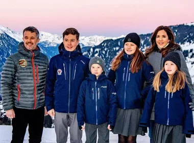 Family in winter jackets poses in snowy mountain landscape during sunset.
