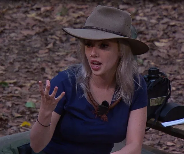 Woman in a blue shirt and hat expressing with her hand in a forest setting.