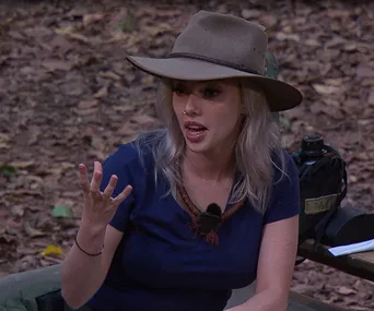 Woman in a blue shirt and hat expressing with her hand in a forest setting.