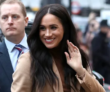 Meghan Markle smiling and waving outside Canada House, wearing a tan coat.