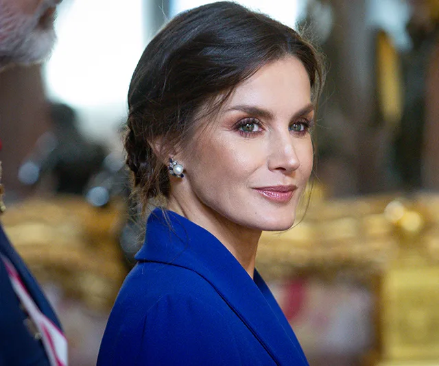 A woman in a blue dress with pearl earrings, looking over her shoulder in an elegant setting.