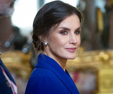 A woman in a blue dress with pearl earrings, looking over her shoulder in an elegant setting.