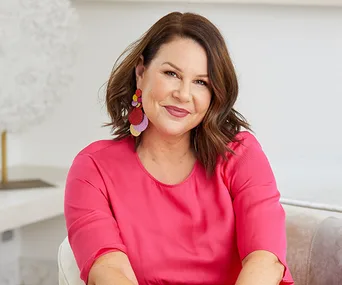 A woman in a pink top sits on a couch, smiling, with colorful earrings and wavy brown hair.