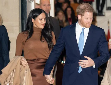 Couple holding hands, dressed formally; woman in brown, man in blue suit, walking past crowd.