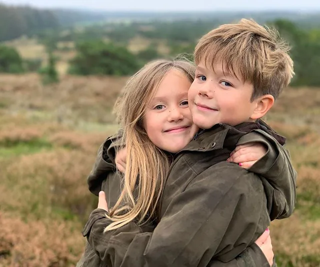 Twins hugging in a grassy, scenic landscape, both wearing dark jackets, with a blurred background of fields and trees.