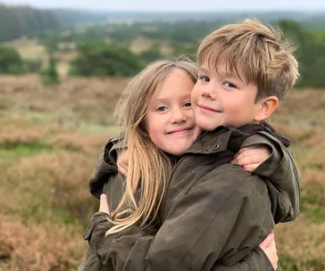 Twins hugging in a grassy, scenic landscape, both wearing dark jackets, with a blurred background of fields and trees.