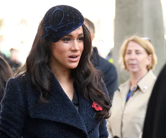 Meghan Markle wearing a dark coat and hat with a red poppy pin during an outdoor event.