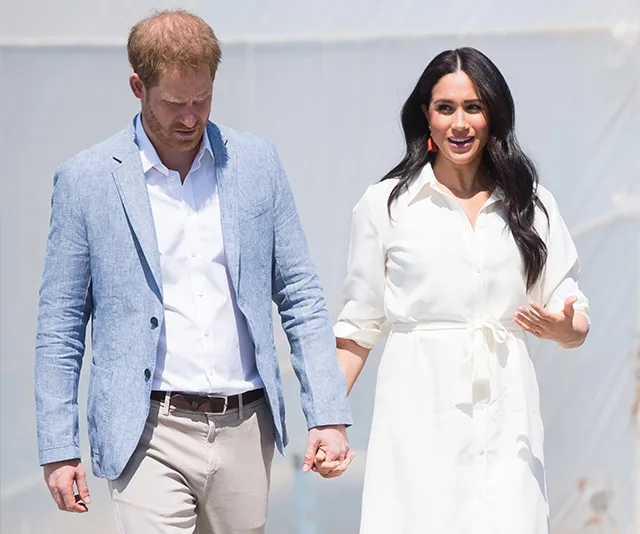 Prince Harry and Meghan Markle walking hand in hand, with Harry in a blue blazer and Meghan in a white dress.