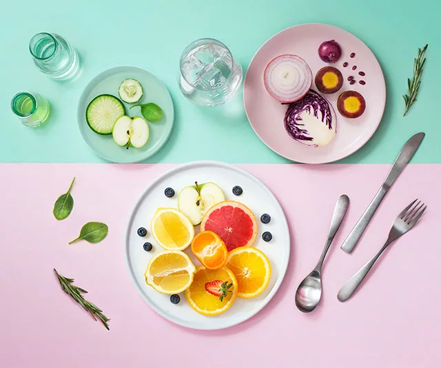Plates of sliced fruits and vegetables on pastel backgrounds with utensils and herbs, illustrating detox diet ingredients.