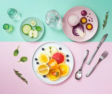 Plates of sliced fruits and vegetables on pastel backgrounds with utensils and herbs, illustrating detox diet ingredients.