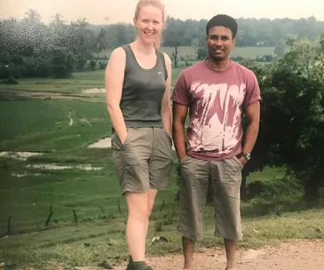 Two people smiling while posing in front of a lush green landscape, wearing casual attire on a sunny day.