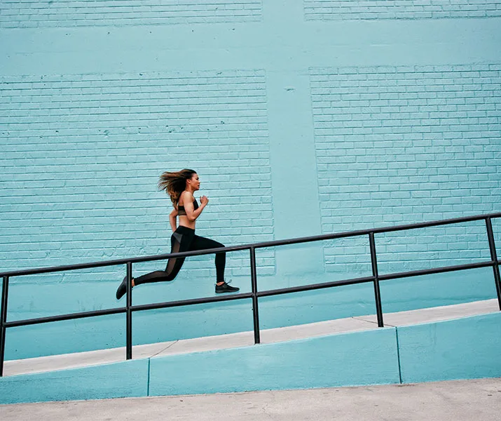 A woman in athletic wear runs uphill beside a turquoise brick wall.