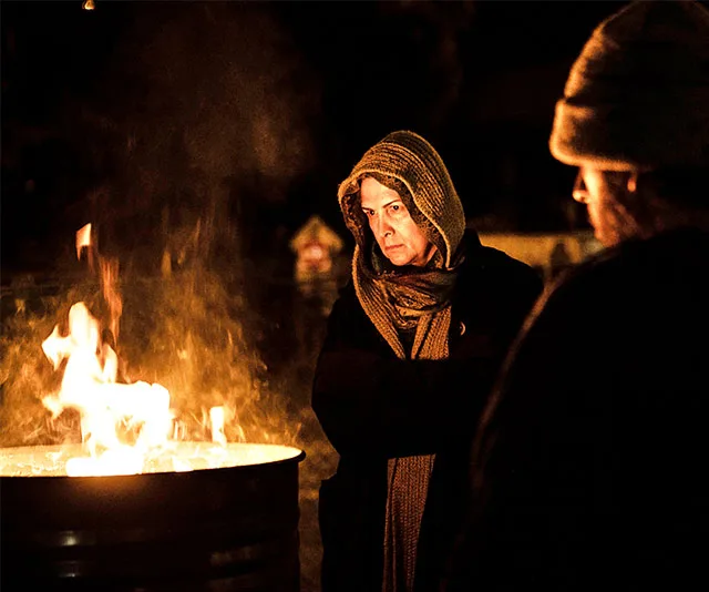 A hooded person stands by a fire barrel at night, looking serious, with another person nearby.