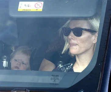Woman in sunglasses driving a car, with a child in the back seat looking slightly upward.