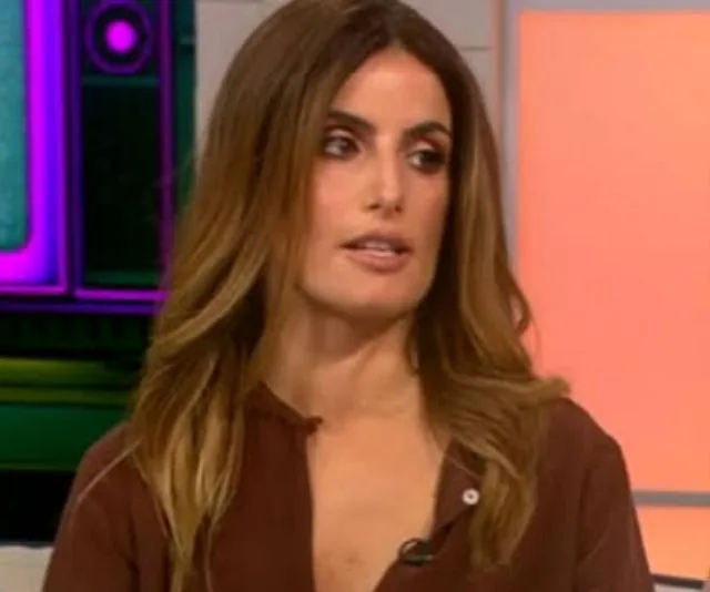 A woman with long brown hair wearing a brown shirt, on a colorful TV set.