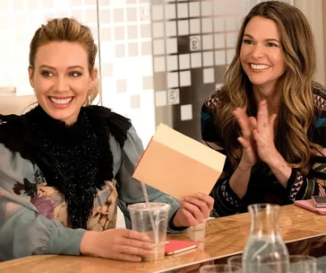 Two women smiling and clapping at a table in an office setting, one holding a file, with a drink nearby.