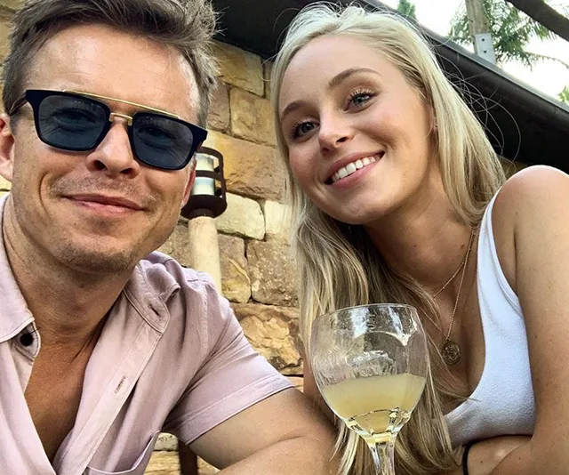 A couple smiles at the camera outdoors; a drink in the foreground.