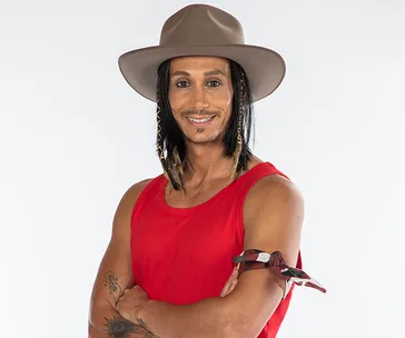 Man in a red sleeveless shirt and hat, smiling with braided hair and a tattoo on his arm, against a plain background.
