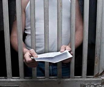 Prisoner holding a handwritten letter behind jail bars, wearing a white tank top.