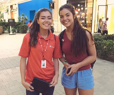Two women who look similar, both smiling, stand together in a mall; one is in a red polo, the other in a red top and blue shorts.