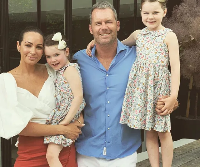 A family of four smiling, two young girls in floral dresses, man in blue shirt, woman in white top, outdoors.