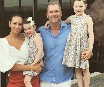 A family of four smiling, two young girls in floral dresses, man in blue shirt, woman in white top, outdoors.