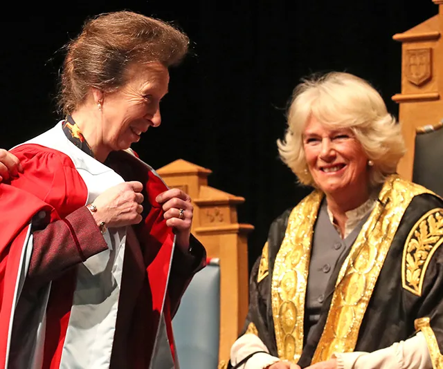 Two women in ceremonial robes, one assisting the other. They're smiling, with ornate chairs in the background.