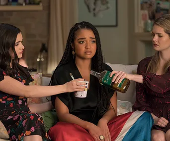 Three women sit on a couch; one looks sad while the others offer ice cream and a drink.