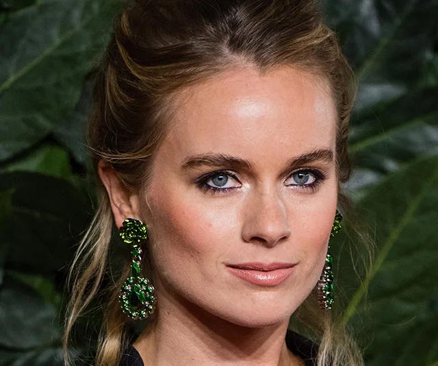 Portrait of a woman with long blonde hair, wearing green jeweled earrings, in front of greenery.