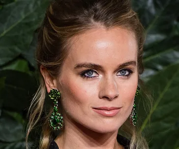 Portrait of a woman with long blonde hair, wearing green jeweled earrings, in front of greenery.
