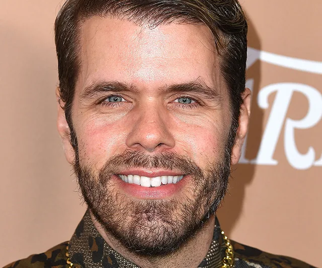 A smiling person with a beard and a patterned shirt at an event with a brown background.