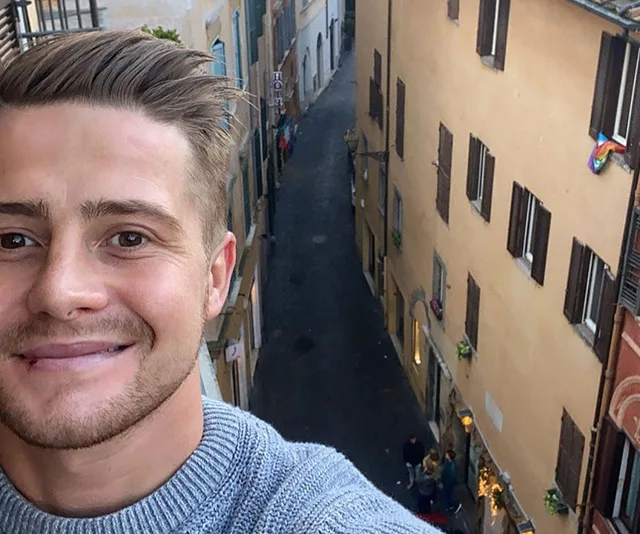 A man takes a selfie from a high point overlooking a narrow European street with colorful buildings and pedestrians.