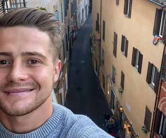 A man takes a selfie from a high point overlooking a narrow European street with colorful buildings and pedestrians.