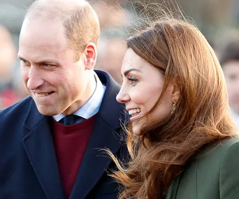Two people are smiling and walking outdoors, nicely dressed, with a crowd in the background.
