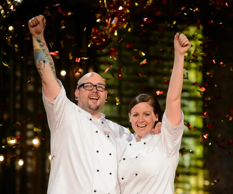 Two chefs celebrate under falling confetti, arms raised in victory, smiling broadly in white uniforms.