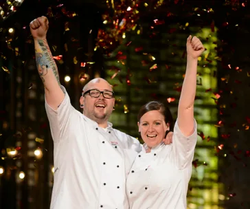 Two chefs celebrate under falling confetti, arms raised in victory, smiling broadly in white uniforms.