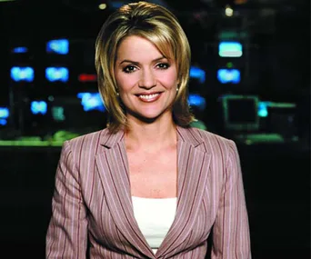 A person smiling in a news studio, wearing a striped blazer with monitors in the background.