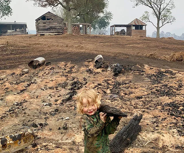 Child holding burnt wood in a scorched landscape with damaged houses after fires, depicting the aftermath of bushfires in Cobargo.