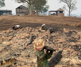 Child holding burnt wood in a scorched landscape with damaged houses after fires, depicting the aftermath of bushfires in Cobargo.