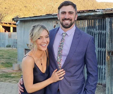 A smiling couple posing outdoors, with the man in a suit and the woman in a dark dress.