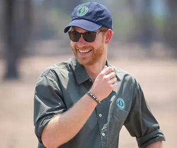 A man in a cap and sunglasses, smiling in an outdoor setting, wearing a shirt with a logo.