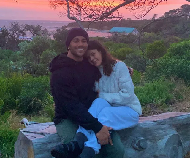 A couple sitting on a wooden bench outdoors, with a sunset and lush greenery in the background.