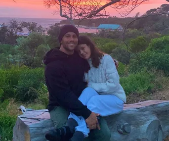 A couple sitting on a wooden bench outdoors, with a sunset and lush greenery in the background.