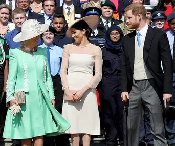 A woman in a green dress and hat stands with a couple in formal attire, surrounded by a diverse group of people.