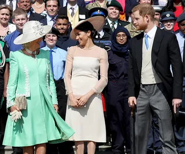 A woman in a green dress and hat stands with a couple in formal attire, surrounded by a diverse group of people.