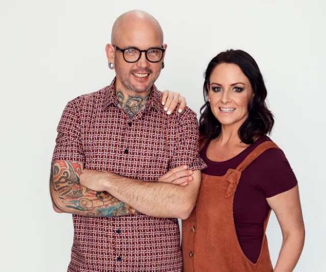 Two people smiling; the person on the left wears glasses and has tattoos, while the person on the right has dark hair.
