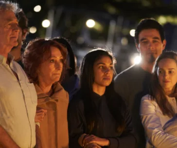 Group of somber individuals gathered outdoors at night, lit by warm ambient light.